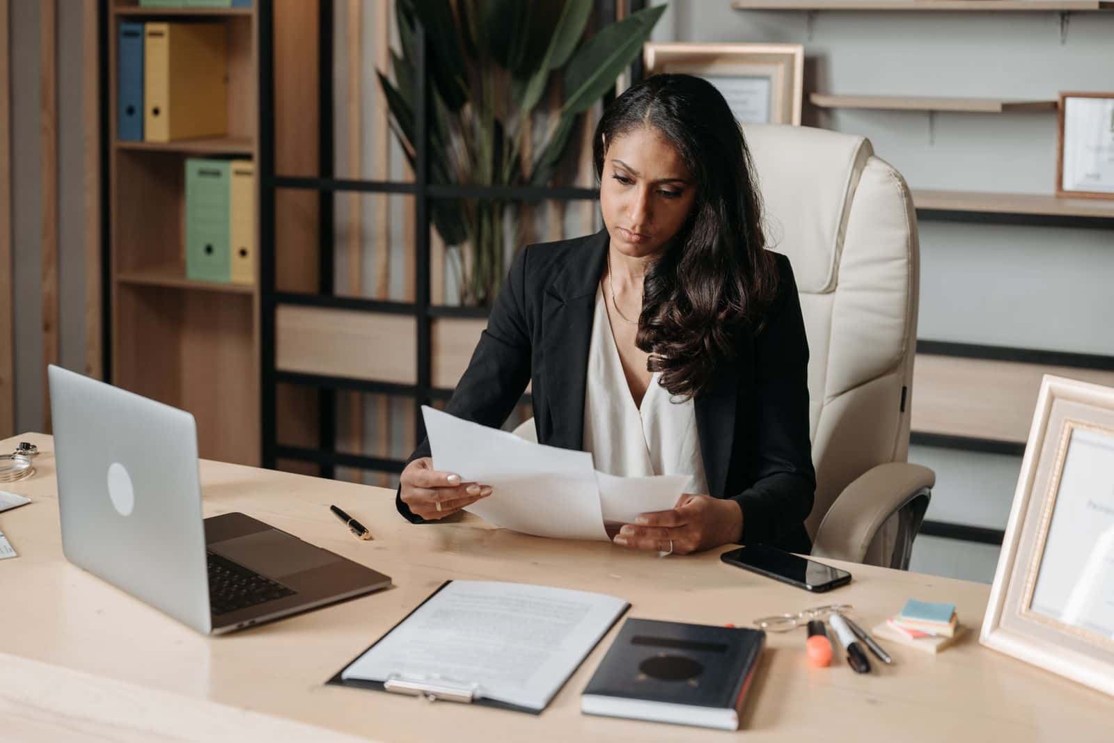 A woman at an office desk reading a document beside a laptop