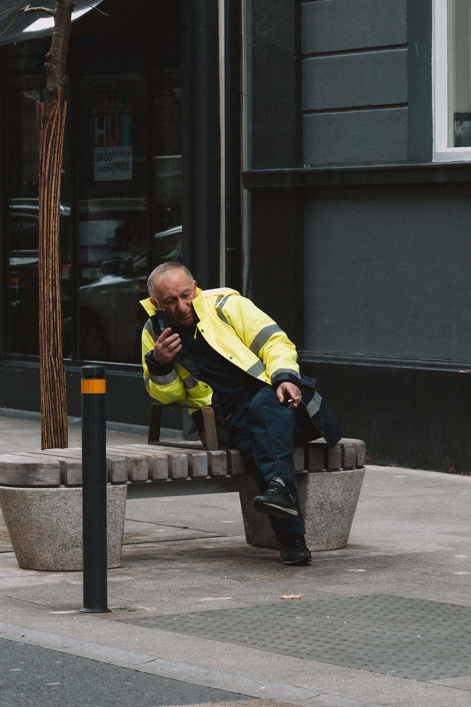 A worker in a hi-vis jacket sitting on a bench outside a building, on the phone
